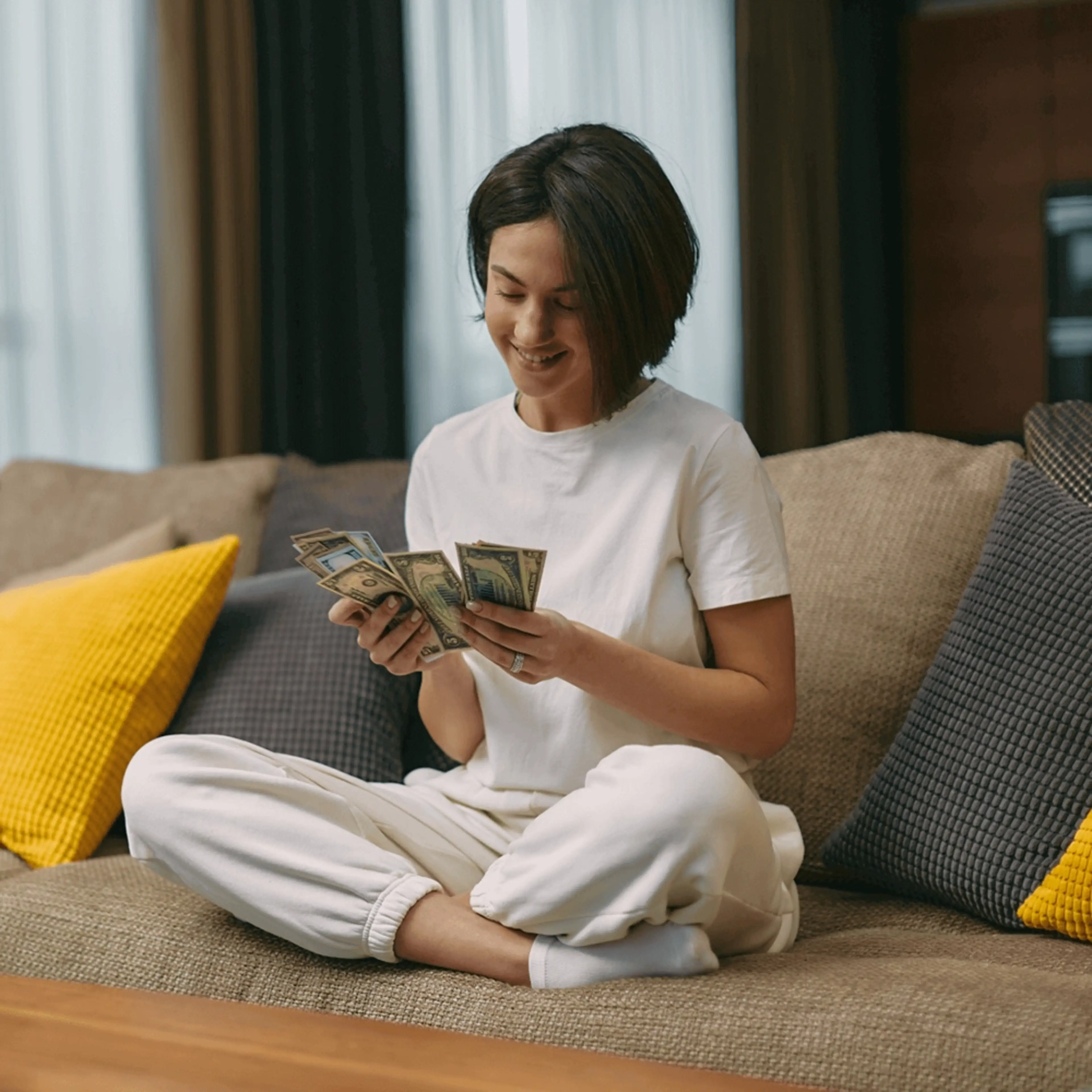 Person in white outfit sitting cross-legged on couch counting money, smiling with yellow pillows nearby.